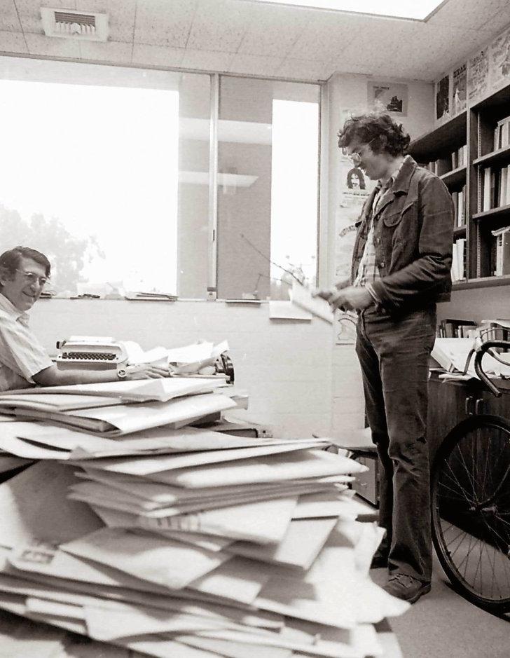 A pile of mail sits in the foreground while two male staff talk in the background